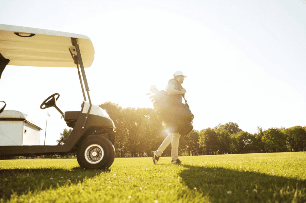 man with a white hat and pants going on a filed and holding a golf bag near the golf cart